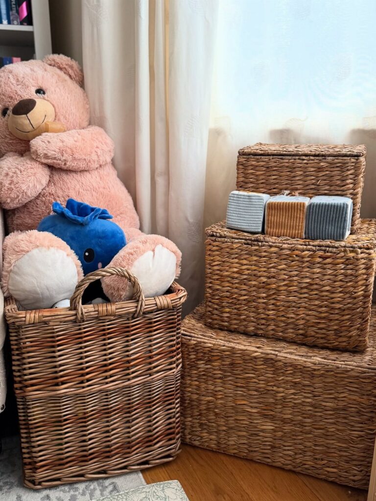 Wicker storage baskets containing a large pink teddy bear and blue plush toy on a wooden floor.