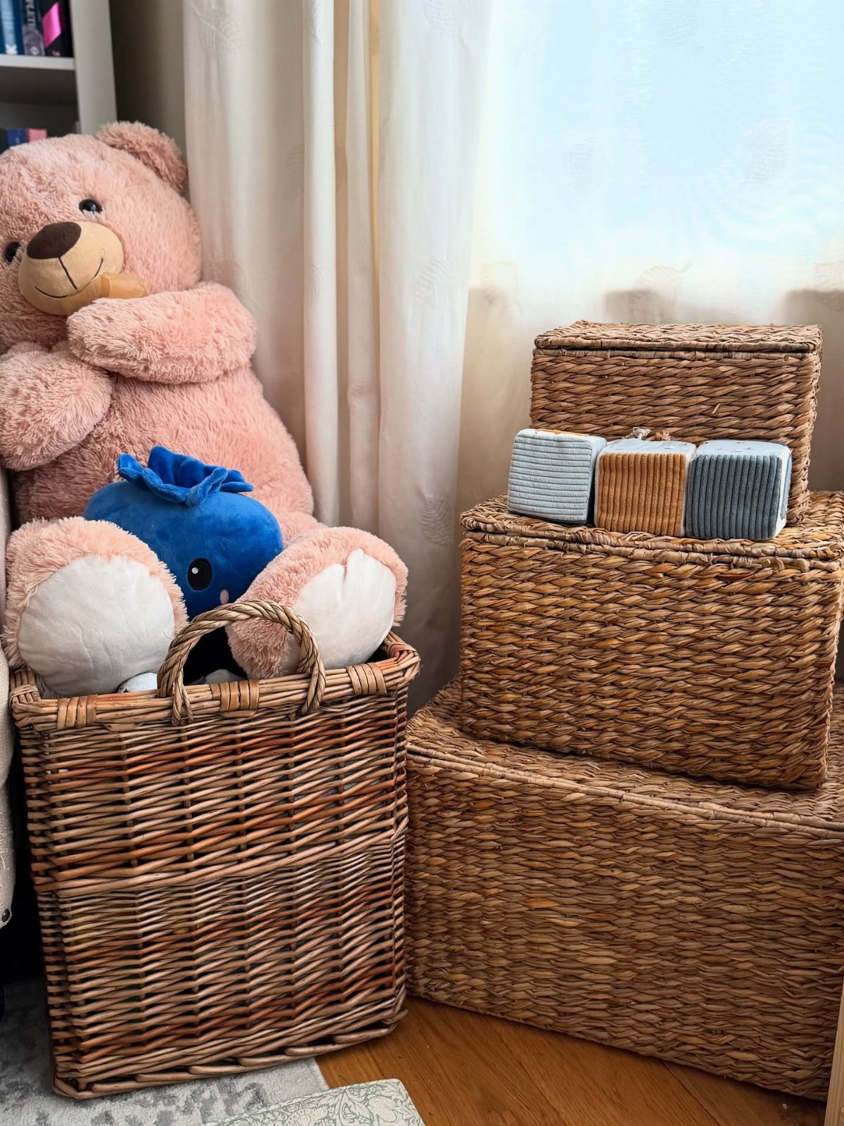 Wicker storage baskets containing a large pink teddy bear and blue plush toy on a wooden floor.