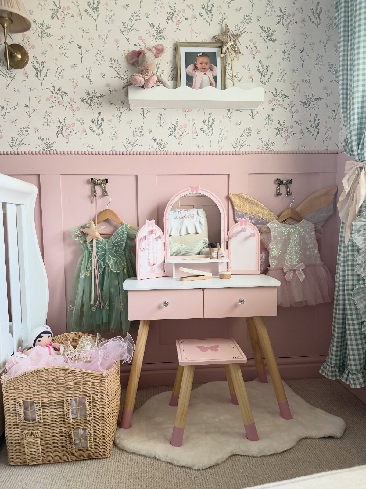 Pink vanity table with costumes hanging on a pink paneled wall in a nursery.
